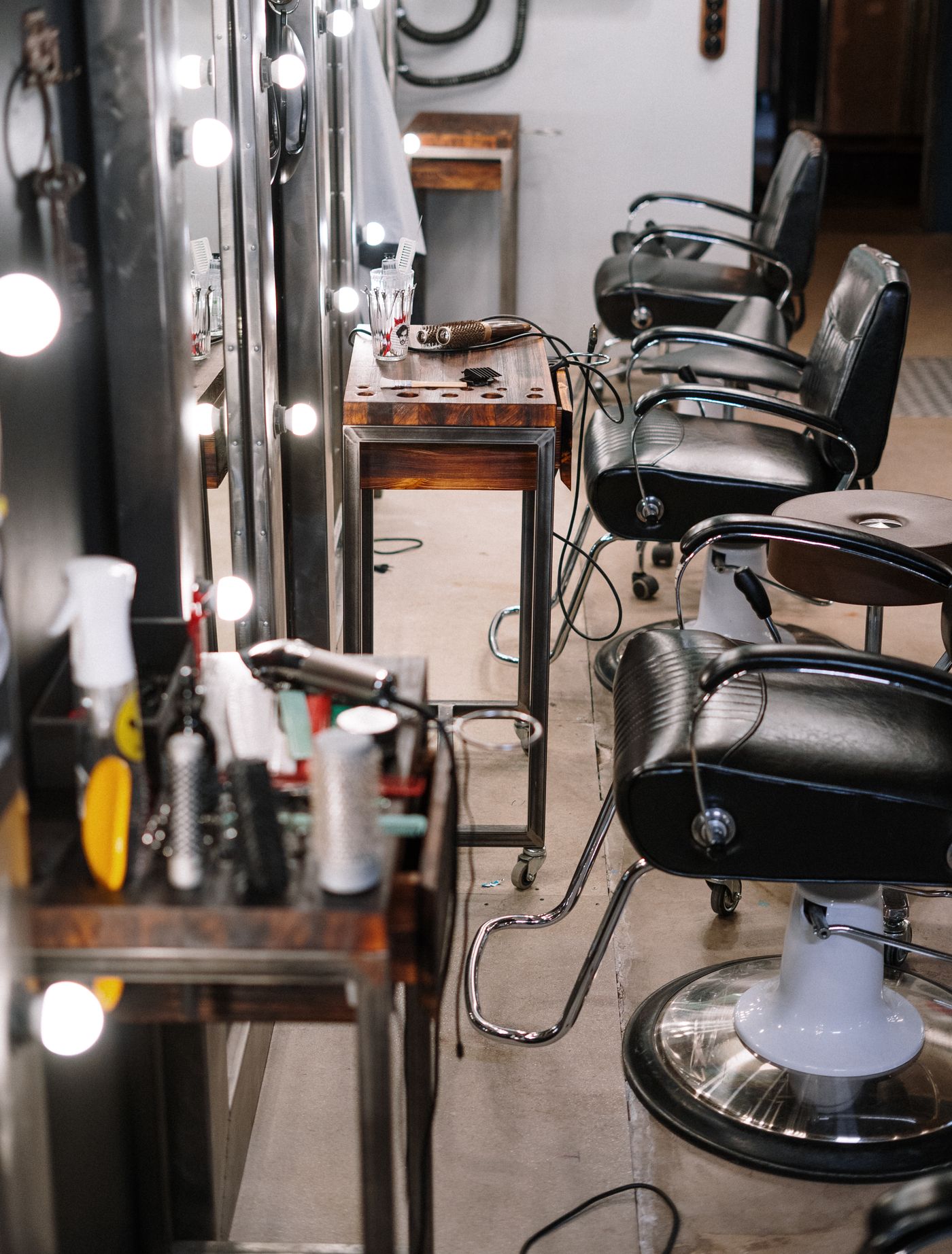 A row of black leather salon chairs beside a bulb-lit mirror, with a trolley holding combs in a glass, brushes and a styling bottle blurred in the foreground.