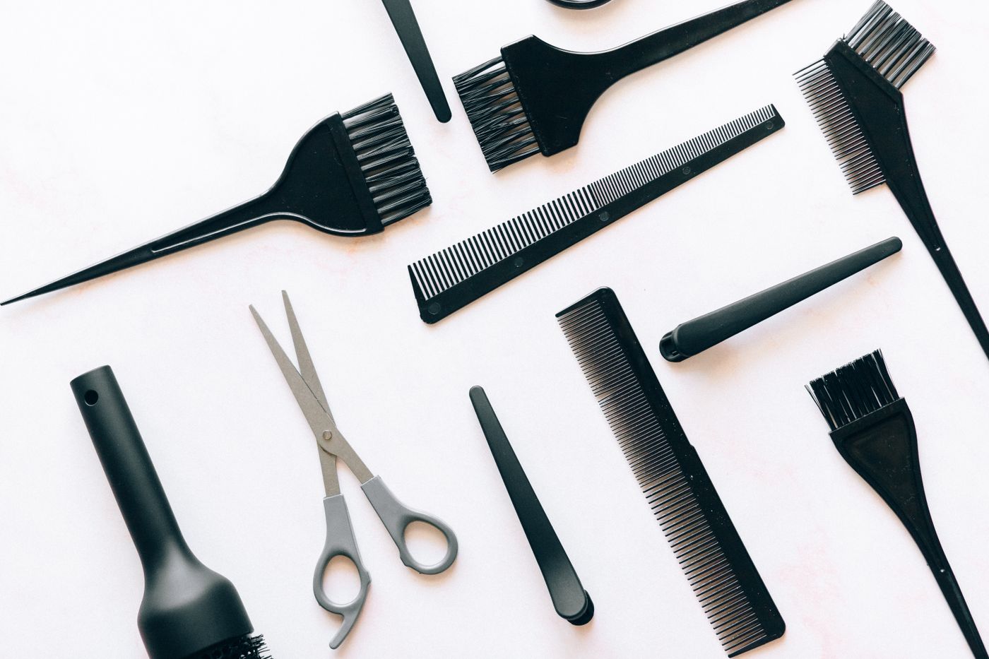 A flat lay of black tint brushes, combs, sectioning clips and grey-handled scissors on a white marble surface.